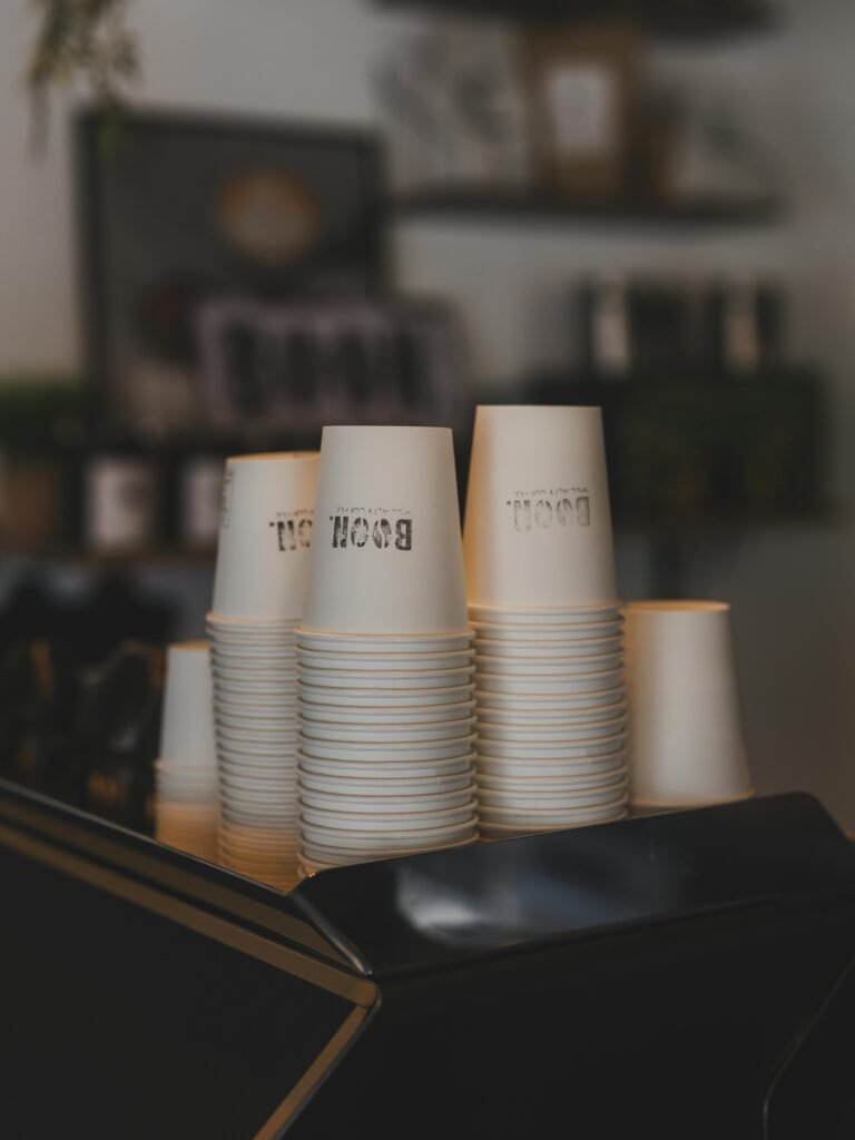 A stack of white paper cups sits on a coffee machine in a warm and cozy café.